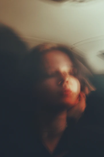 A softly lit, intimate portrait of the artist surrounded by subtle, warm-toned abstract shapes.