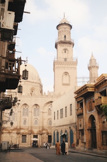 A street scene featuring historical architecture, with a prominent mosque showcasing a large dome and a tall minaret. The buildings display intricate designs and arched windows. A few people dressed in traditional attire walk along the cobblestone street, lending the scene a cultural and historical ambiance.