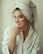 A person wearing a white bathrobe with a towel wrapped around their head, smiling gently with their hand near the face, showcasing intricate nail art.
