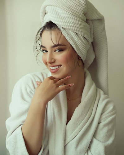 A person wearing a white bathrobe with a towel wrapped around their head, smiling gently with their hand near the face, showcasing intricate nail art.