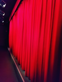 Bright red theater curtains are drawn across a stage, illuminated by stage lights from above. The curtain fabric appears heavy and lush, suggesting a formal or dramatic setting.