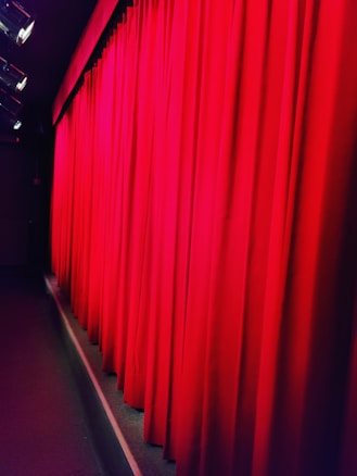 Bright red theater curtains are drawn across a stage, illuminated by stage lights from above. The curtain fabric appears heavy and lush, suggesting a formal or dramatic setting.