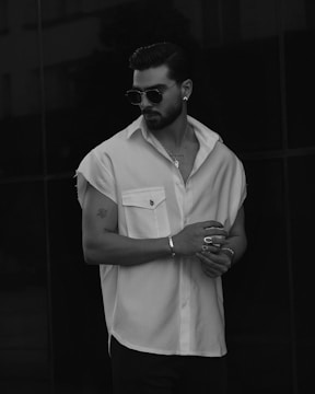 A man with a stylish beard and slicked-back hair is wearing dark sunglasses and a button-up shirt with short sleeves, posing against a dark background. He is accessorized with rings, earrings, and a bracelet.