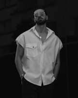 A person stands confidently wearing sunglasses and a sleeveless shirt with tattoos visible on their arms. The monochromatic style adds a modern and stylish vibe.