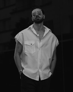 A person stands confidently wearing sunglasses and a sleeveless shirt with tattoos visible on their arms. The monochromatic style adds a modern and stylish vibe.