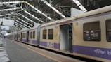 A train stands stationary at an indoor railway platform with large metal beams supporting the roof. The train car doors are open, revealing an empty interior. The platform is mostly empty, with some metal benches arranged in rows alongside the track.