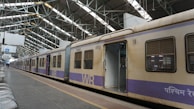 A train stands stationary at an indoor railway platform with large metal beams supporting the roof. The train car doors are open, revealing an empty interior. The platform is mostly empty, with some metal benches arranged in rows alongside the track.