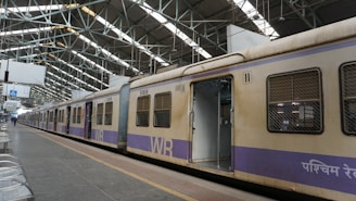 A train stands stationary at an indoor railway platform with large metal beams supporting the roof. The train car doors are open, revealing an empty interior. The platform is mostly empty, with some metal benches arranged in rows alongside the track.