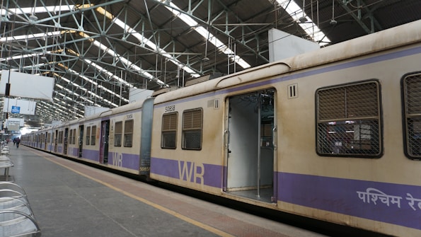 A train stands stationary at an indoor railway platform with large metal beams supporting the roof. The train car doors are open, revealing an empty interior. The platform is mostly empty, with some metal benches arranged in rows alongside the track.