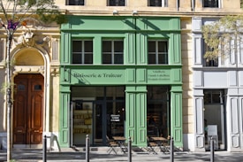 A charming street scene featuring a green-painted storefront labeled 'Rôtisserie & Traiteur' situated between two historic-style buildings. The facade includes large windows and an entrance with small tables and chairs set outside. A decorative stone doorway with intricate carvings is prominent on the left, and a tree with light green leaves is in the foreground.