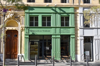 A charming street scene featuring a green-painted storefront labeled 'Rôtisserie & Traiteur' situated between two historic-style buildings. The facade includes large windows and an entrance with small tables and chairs set outside. A decorative stone doorway with intricate carvings is prominent on the left, and a tree with light green leaves is in the foreground.