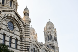 A detailed view of a large stone building with intricate architecture, featuring striped patterns and multiple domed towers topped with crosses. The structure showcases Romanesque and Byzantine design elements, with arched windows and decorative stonework.