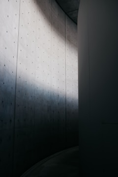 A minimalist architectural scene featuring a tall, curved concrete wall with a series of small, evenly spaced holes. The play of light and shadow creates a gradient across the surface, adding depth to the monochrome color palette.