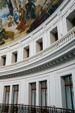 A grand interior view with a curved wall featuring large windows with wooden frames. Above, a detailed fresco depicts a historical scene with people engaged in various activities, including farming and trading, set against a rustic backdrop. The architecture includes ornate moldings and a decorative metal railing.