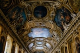 An ornate ceiling featuring intricate paintings and elaborate architectural decorations. The artwork includes detailed figures and scenes surrounded by lavish gold embellishments, enhancing the opulence and grandeur of the space.