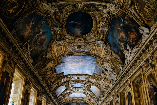 An ornate ceiling featuring intricate paintings and elaborate architectural decorations. The artwork includes detailed figures and scenes surrounded by lavish gold embellishments, enhancing the opulence and grandeur of the space.