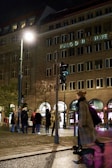 A nighttime urban scene with a group of people standing on a wet street corner near an old building with illuminated windows. The building facade has the words 'Haus der Schweiz' displayed. A bright streetlight casts a glow over the area. Several pedestrians are visible, including a person in motion wearing a coat.