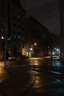 Dimly lit urban street with wet pavement reflecting streetlights. Tall buildings line the street, and a single person is visible walking along the sidewalk. The scene is quiet and appears to be set at night.