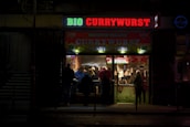 A small illuminated fast-food restaurant is open in a dark setting, with a bright sign reading 'BIO CURRYWURST'. Several people are gathered inside, likely ordering or eating food, and the interior has a welcoming, warm glow.