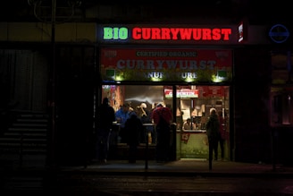 A small illuminated fast-food restaurant is open in a dark setting, with a bright sign reading 'BIO CURRYWURST'. Several people are gathered inside, likely ordering or eating food, and the interior has a welcoming, warm glow.