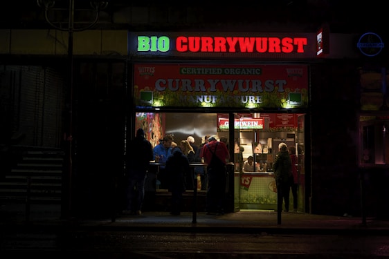 A small illuminated fast-food restaurant is open in a dark setting, with a bright sign reading 'BIO CURRYWURST'. Several people are gathered inside, likely ordering or eating food, and the interior has a welcoming, warm glow.