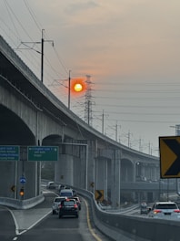 A highway curves under an elevated railway track, with multiple cars traveling on it. Overhead, a large transmission tower stands against a hazy sky with a vibrant orange sun visible near the horizon. Directional signs are present, indicating destinations like Jati Asih and Bogor. The scene is framed by concrete structures and power lines.