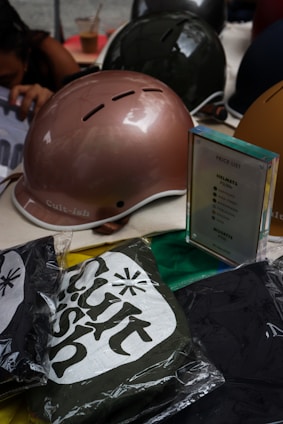 A collection of helmets in metallic brown and dark green, neatly arranged alongside a clear price list display. In front of the helmets, several packaged items with the 'Cult-ish' logo are visible, possibly clothing. A person is partially visible in the background, reaching for something. An iced coffee cup is seen behind the helmets.