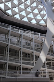 A modern architectural structure with multiple levels of open balconies and railings, illuminated by a large geometric glass roof with a grid-like pattern. The structure is primarily composed of white and gray materials. A single person is visible on one of the lower balconies, adding a sense of scale to the space.
