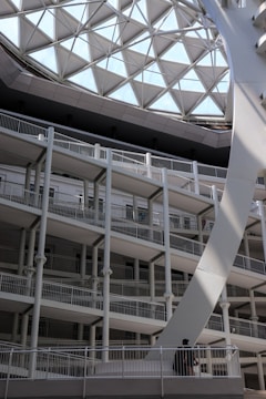 A modern architectural structure with multiple levels of open balconies and railings, illuminated by a large geometric glass roof with a grid-like pattern. The structure is primarily composed of white and gray materials. A single person is visible on one of the lower balconies, adding a sense of scale to the space.