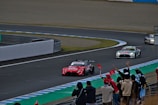A group of race cars speeds around a track, with spectators watching and cheering from the sidelines. The lead car is red and prominently displays sponsorship logos, while several other cars follow closely behind. The track is surrounded by safety barriers and features colorful painted sections on the pavement. A crowd of people, some with flags, line the edge of the track, creating an atmosphere of excitement and action.