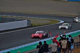 A group of race cars speeds around a track, with spectators watching and cheering from the sidelines. The lead car is red and prominently displays sponsorship logos, while several other cars follow closely behind. The track is surrounded by safety barriers and features colorful painted sections on the pavement. A crowd of people, some with flags, line the edge of the track, creating an atmosphere of excitement and action.