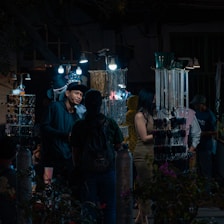 A nighttime market scene with several people browsing through a variety of accessories displayed on illuminated stands. The market stalls are adorned with hanging items such as necklaces and bracelets, lit by bright bulbs, creating a contrast with the dark surroundings. One of the shoppers is engaged in conversation with a vendor.