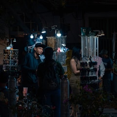 A nighttime market scene with several people browsing through a variety of accessories displayed on illuminated stands. The market stalls are adorned with hanging items such as necklaces and bracelets, lit by bright bulbs, creating a contrast with the dark surroundings. One of the shoppers is engaged in conversation with a vendor.