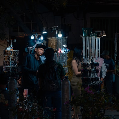 A nighttime market scene with several people browsing through a variety of accessories displayed on illuminated stands. The market stalls are adorned with hanging items such as necklaces and bracelets, lit by bright bulbs, creating a contrast with the dark surroundings. One of the shoppers is engaged in conversation with a vendor.