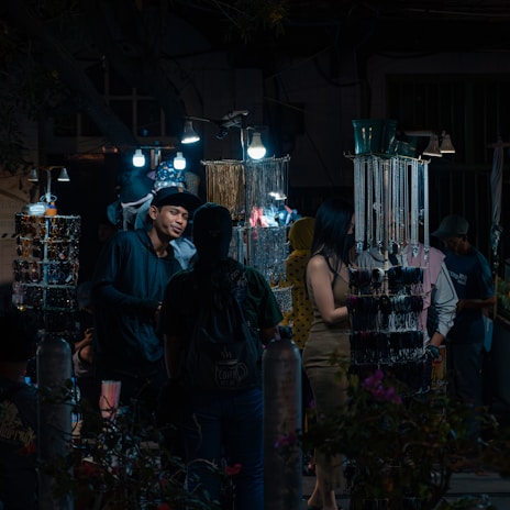 A nighttime market scene with several people browsing through a variety of accessories displayed on illuminated stands. The market stalls are adorned with hanging items such as necklaces and bracelets, lit by bright bulbs, creating a contrast with the dark surroundings. One of the shoppers is engaged in conversation with a vendor.