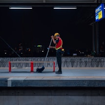 A person wearing a safety vest and helmet is cleaning a train platform with a broom at night. The platform is dimly lit, with a city skyline visible in the background. Red barriers are placed around the platform area, and a sign board can be seen overhead.