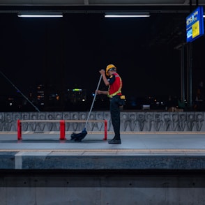 A person wearing a safety vest and helmet is cleaning a train platform with a broom at night. The platform is dimly lit, with a city skyline visible in the background. Red barriers are placed around the platform area, and a sign board can be seen overhead.