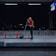 A person wearing a safety vest and helmet is cleaning a train platform with a broom at night. The platform is dimly lit, with a city skyline visible in the background. Red barriers are placed around the platform area, and a sign board can be seen overhead.