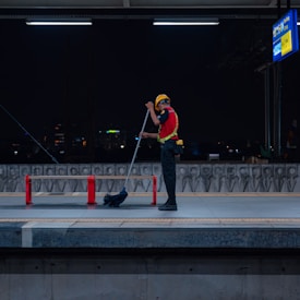 A person wearing a safety vest and helmet is cleaning a train platform with a broom at night. The platform is dimly lit, with a city skyline visible in the background. Red barriers are placed around the platform area, and a sign board can be seen overhead.