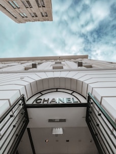 A low-angle view of a building with a prominent 'CHANEL' sign above a gated entrance. The facade of the building is white with architectural detailing, and the sky above is filled with soft clouds.