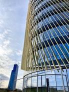 A modern building with a dynamic facade featuring vertical, angled slats and reflective glass panels. In the background, a tall skyscraper with a crane is visible, suggesting construction work. The sky is partly cloudy, creating a serene urban setting.