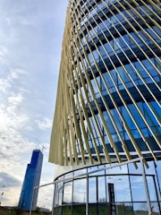 A modern building with a dynamic facade featuring vertical, angled slats and reflective glass panels. In the background, a tall skyscraper with a crane is visible, suggesting construction work. The sky is partly cloudy, creating a serene urban setting.