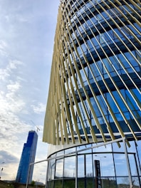 A modern building with a dynamic facade featuring vertical, angled slats and reflective glass panels. In the background, a tall skyscraper with a crane is visible, suggesting construction work. The sky is partly cloudy, creating a serene urban setting.