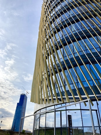 A modern building with a dynamic facade featuring vertical, angled slats and reflective glass panels. In the background, a tall skyscraper with a crane is visible, suggesting construction work. The sky is partly cloudy, creating a serene urban setting.