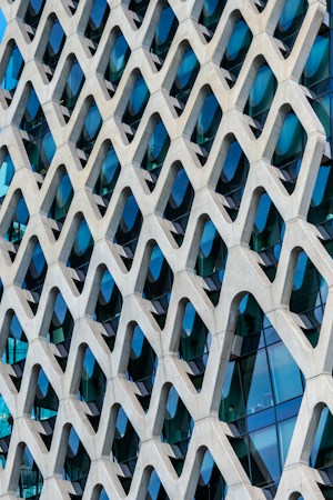 A modern architectural facade featuring a geometric pattern of repeated Y-shaped structures. The design creates a three-dimensional effect with alternating shadows and reflections. Behind the pattern, reflective glass windows are visible, contributing to a sleek and contemporary aesthetic.