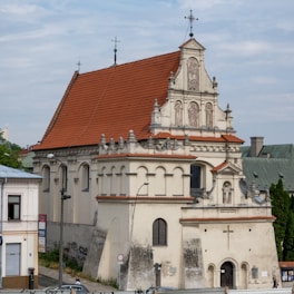 A historic church building with intricate architectural features and a prominent red-tiled roof. The facade includes religious sculptures and arched windows, and the structure is situated in an urban environment with nearby buildings and street signs.