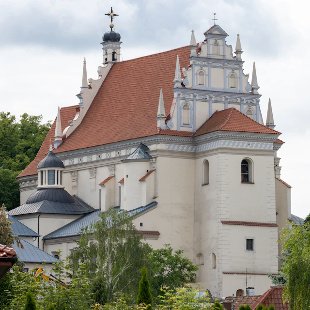 A white Baroque church with a steep red tile roof, scalloped pinnacled gable, and an octagonal side chapel dome.