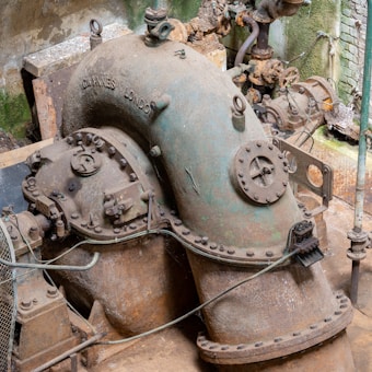 An industrial setting with a large, rusted metal pipe structure, possibly part of an old machinery system. The pipe has numerous bolts and valves, with some signs of corrosion and wear. The surrounding area is gritty with concrete and metal elements, partially weathered with hints of green patina and moss on the walls and machinery.