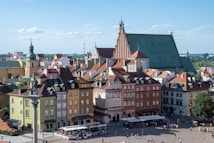 A historic city square with a variety of charming and colorful buildings featuring ornate architecture. The scene includes a large church with a green roof and spire. People are walking around the cobblestone streets, and outdoor seating is visible in the square. The image is taken during the day, with a clear blue sky.