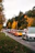 A lineup of luxury sports cars is parked along a narrow road. The cars are in various colors, including white, orange, red, and black, and are positioned in a single file. The scene is set during autumn with vibrant foliage, as the trees along the road display a mix of green and golden-yellow leaves.